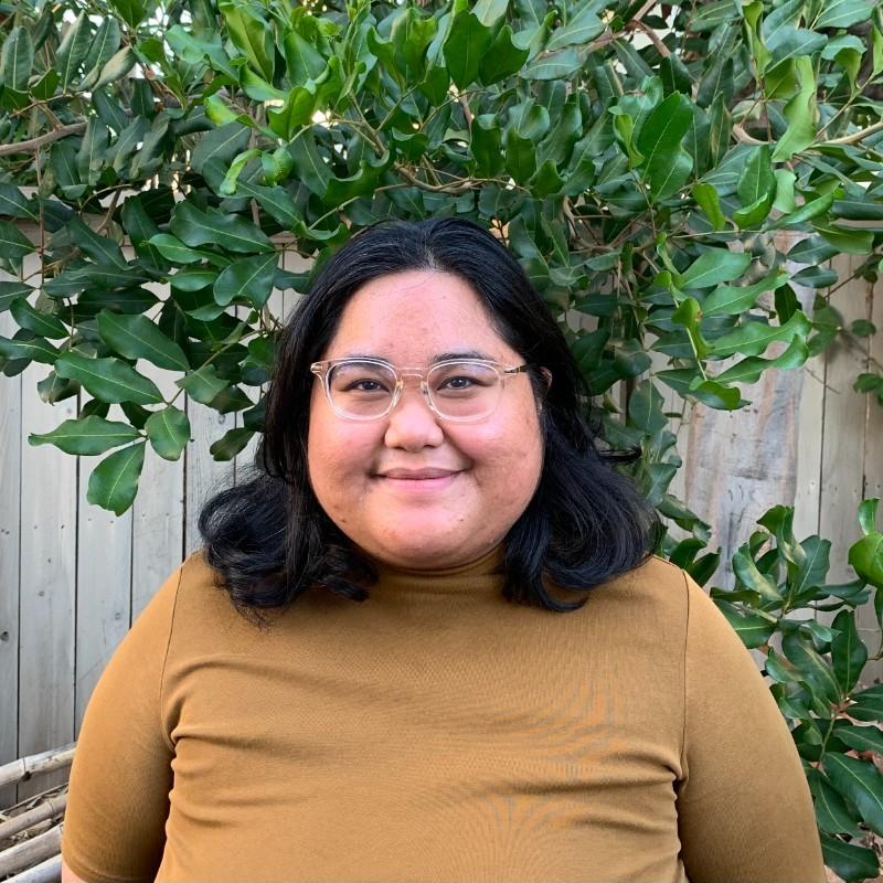 A picture of Andrea standing in front of a tree. Andrea has brown skin and shoulder-length, black hair. She is wearing square eyeglasses and is smiling at the camera.
