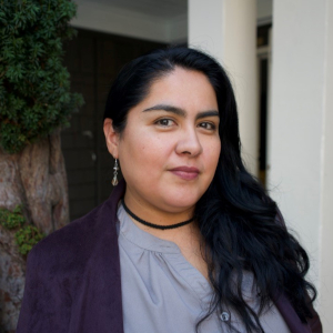Closeup image of Aurora Alvarado whose hair is parted to the side hanging loosely over their left shoulder. Aurora is smiling, and wearing a lavender color blouse, and purple blazer.