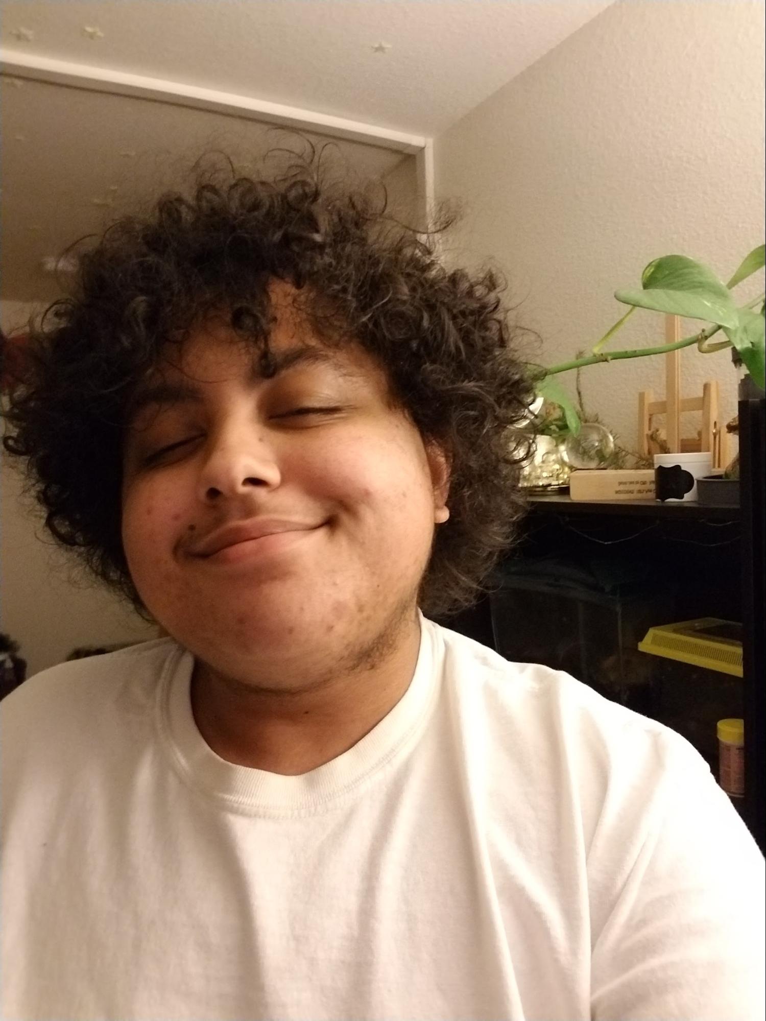 A black, androgynous, young man smiles at the camera. He's wearing a basic white t-shirt in a lightly decorated room, with a plant hanging behind him.
