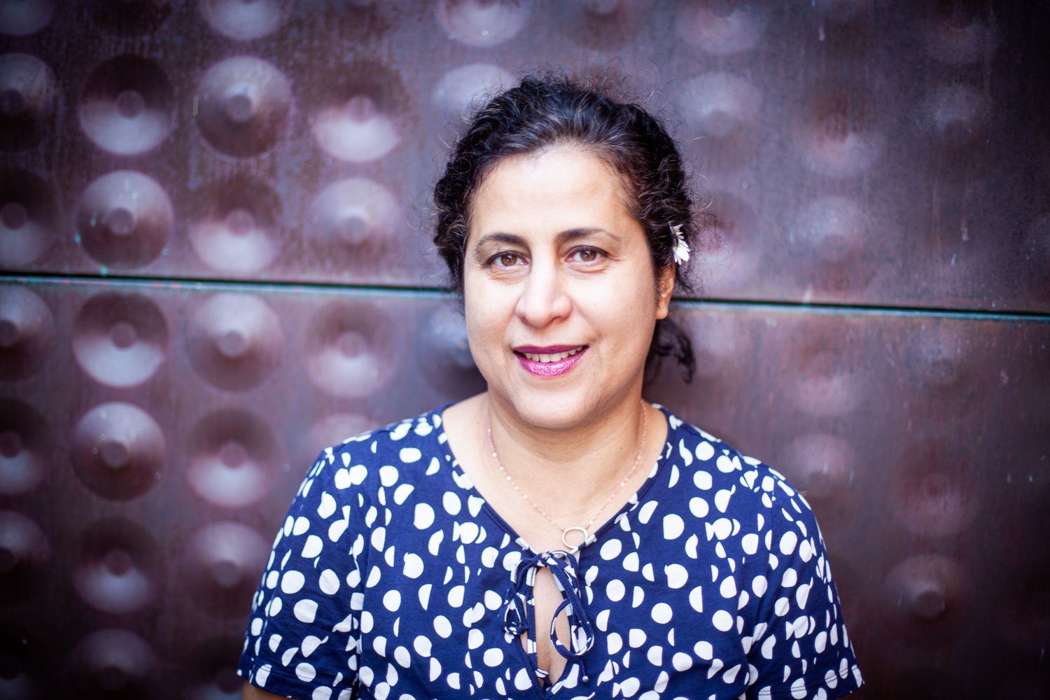 Orkid Sassouni smiling at a camera, standing against the cooper-made wall, female presenting with brown-olive skin, in late 40s, wearing navy shirt with cream color spots. Hair buns back with one white flower placed behind the ear.