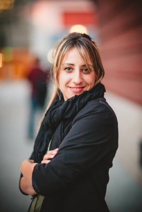 Image of Cecile Puretz, a white woman standing with her arms crossed in front of a brick wall and street. She has brown hair with bangs, is wearing a black blazer and scarf, and is smiling at the camera.